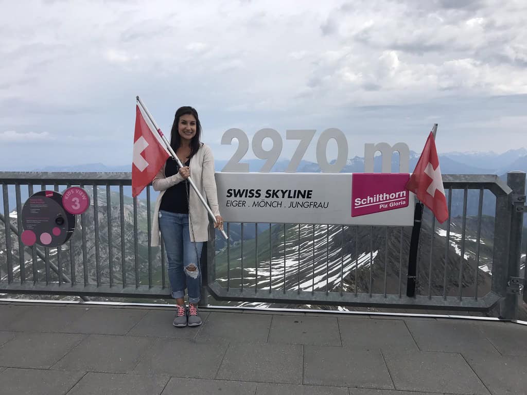 Swiss Skyline viewpoint with woman holding Swiss flags, overlooking mountains and clouds.