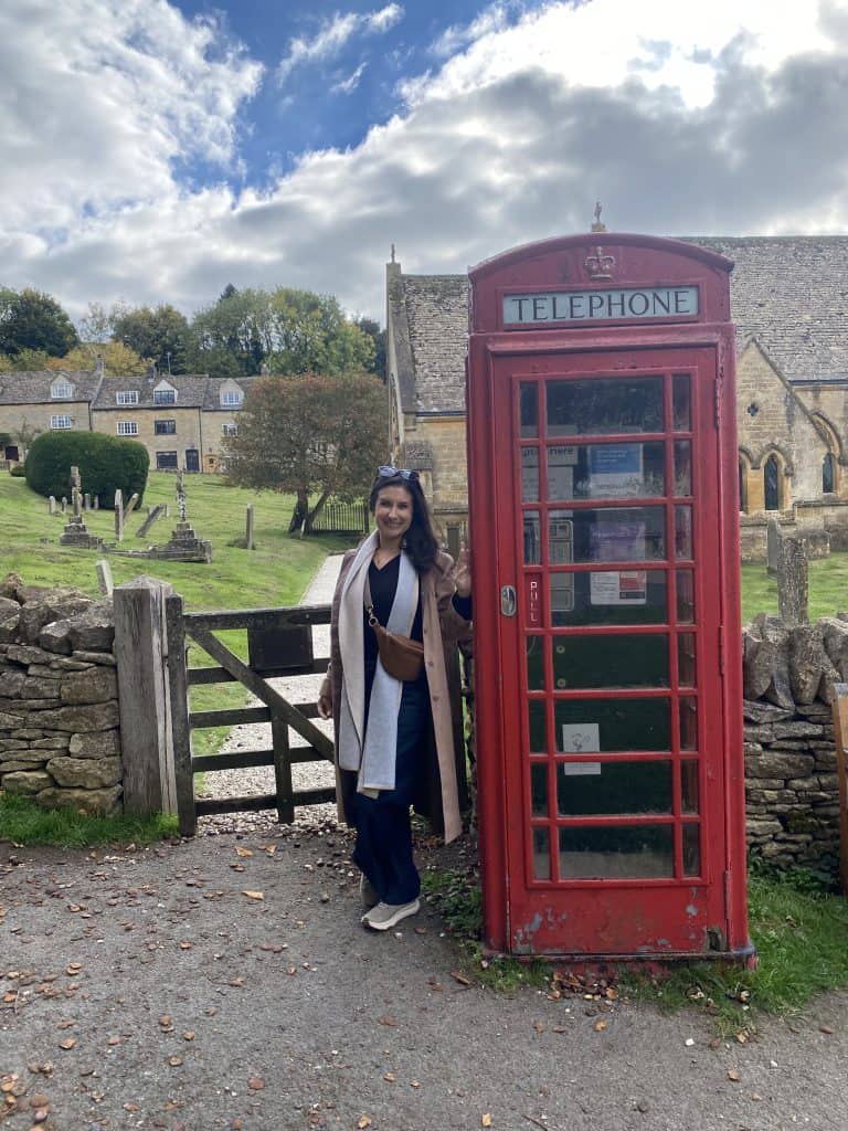 Woman standing next to a classic red British telephone booth in a scenic village setting.